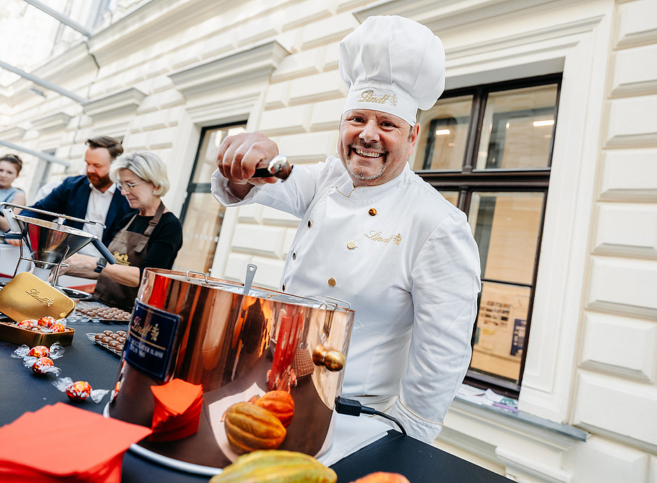 Chocolatier mit weißer Kochhaube präsentiert lächelnd eine kleine Lindt-Schokokugel ©© Nadja Fuchs 