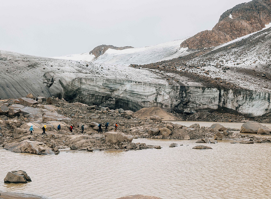 Eine Gruppe von wandernden Menschen, die am Ufer vor einer Eiswand auf einem Gletscher entlanggehen, mit Wasser und Felsen ©Karl Steinegger