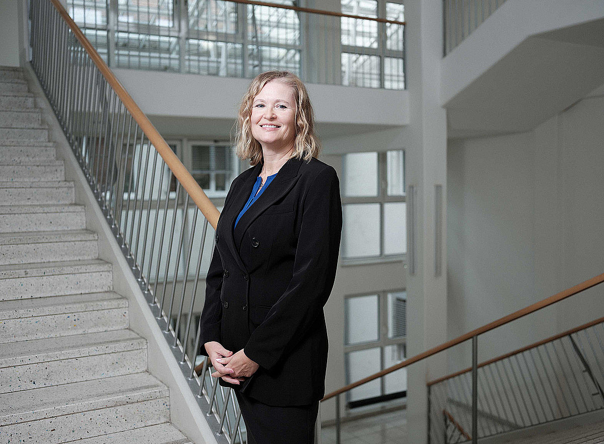 eine Frau mit schulterlangen blonden Haaren, die einen schwarzen Anzug trägt und lächelnd in einem Stiegenhaus steht ©Uni Graz/Schaffernak