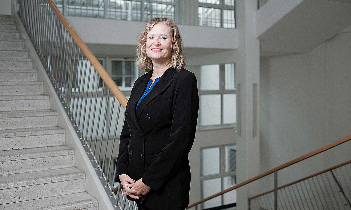 eine Frau mit schulterlangen blonden Haaren, die einen schwarzen Anzug trägt und lächelnd in einem Stiegenhaus steht