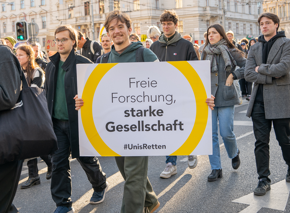 Junger Mann trägt bei einer Demonstration auf der Straße ein Schild mit der Aufschrift „Freie Forschung, starke Gesellschaft #UnisRetten“. 