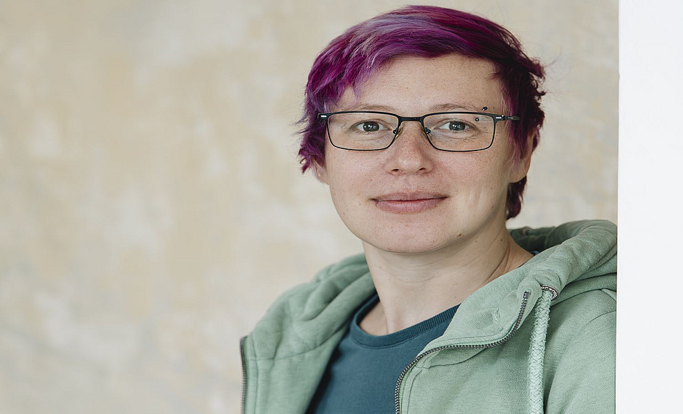 Porträt einer Frau mit kurzen violetten Haaren, die eine Brille und einen grünen Kapuzenpullover trägt und vor einer beigen Wand steht und in die Kamera lächelt.