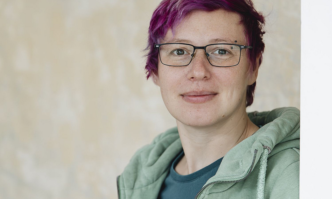 Porträt einer Frau mit kurzen violetten Haaren, die eine Brille und einen grünen Kapuzenpullover trägt und vor einer beigen Wand steht und in die Kamera lächelt.