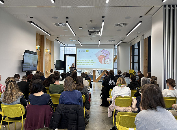 eine Frau steht vor einem Tisch mit Laptop und hält einen Vortrag, hinter ihr sieht man die Leinwand mit der Präsentation. Im Publikum sitzen viele Menschen und hören zu ©Uni Graz/Schmieder