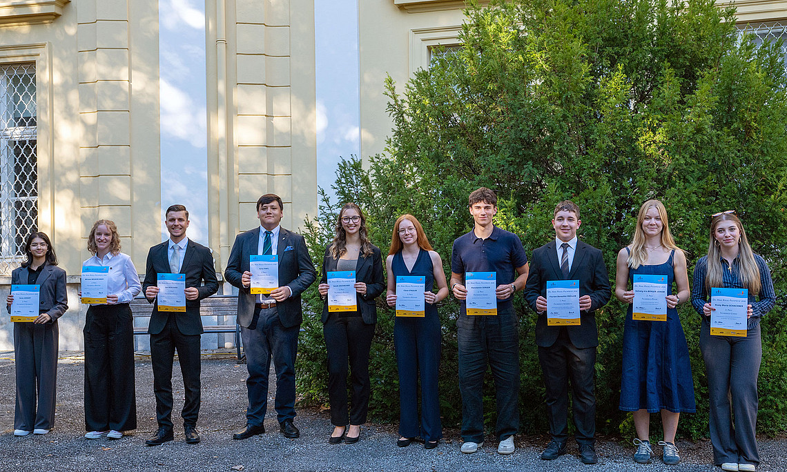 Ten young adults are standing side by side in front of a building, mostly dressed in formal attire and holding certificates in their hands.
