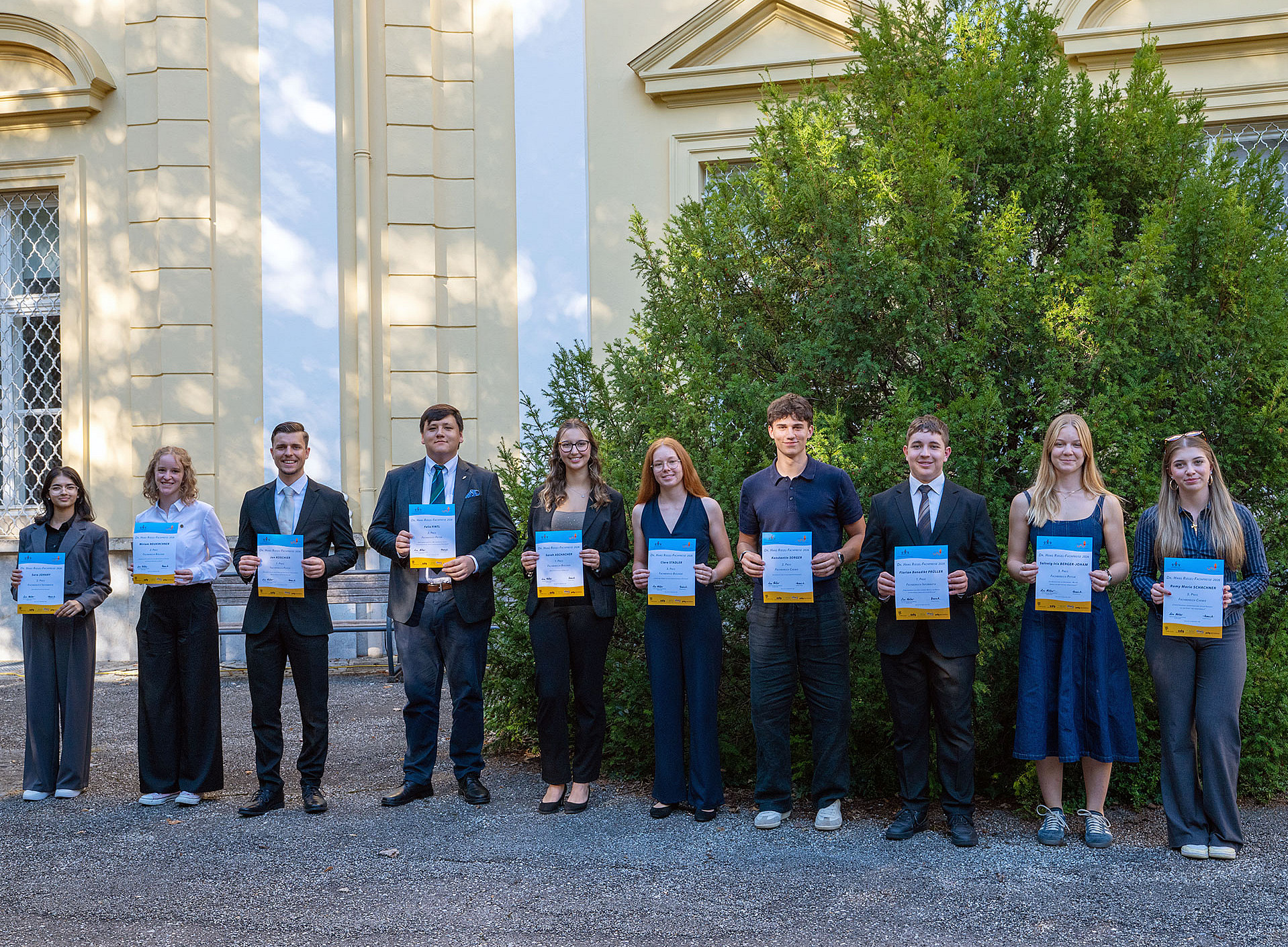 Ten young adults are standing side by side in front of a building, mostly dressed in formal attire and holding certificates in their hands. ©Uni Graz/Weber