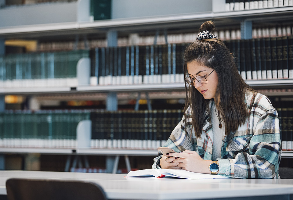Eine Studentin sitzt in der Universitätsbibliothek mit aufgeschlagenem Buch an einem Tisch vor einem großen Bücherregal und sieht auf ihr Handy.