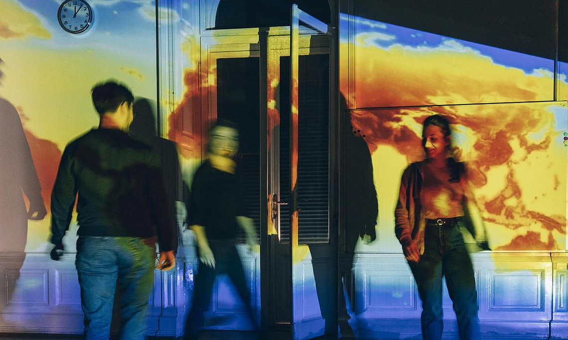 Mehrere Personen schauen sich eine Ausstellung an. Ein Bild wird mithilfe eines Projektors auf die Wand projiziert, was zu einer stimmungsvollen Atmosphäre beiträgt.