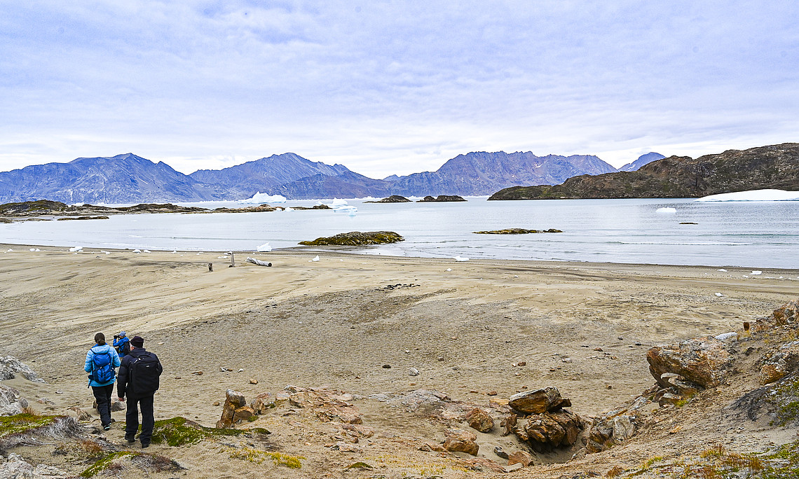 Drei Personen wandern auf einem Sandstrand, im Hintergrund das Meer und Berge