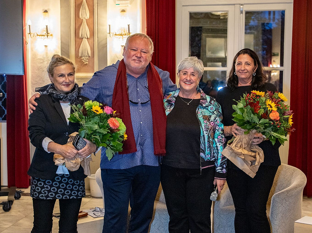 Drei Frauen und ein Mann, die aufgereiht nebeneinander stehen. Die beiden Frauen links und rechts außen halten Blumensträuße, alle lächeln in die Kamera. 