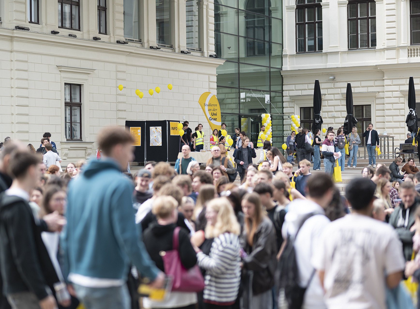 Eine große Gruppe von Menschen, überwiegend junge Erwachsene, steht auf einem belebten Platz vor einem historischen Universitätsgebäude mit heller Fassade und großen Fenstern. ©Uni Graz/Angele
