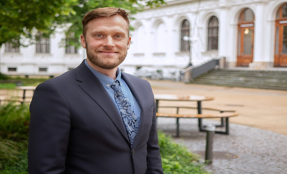 Person im Anzug mit gemusterter Krawatte steht in einem Hof mit Bänken und Grünflächen vor einem historischen Gebäude mit Arkaden.