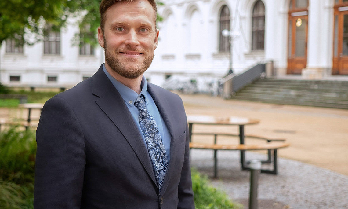 Person in a suit and a patterned tie is standing in a courtyard with benches and green spaces in front of a historic building with arcades.