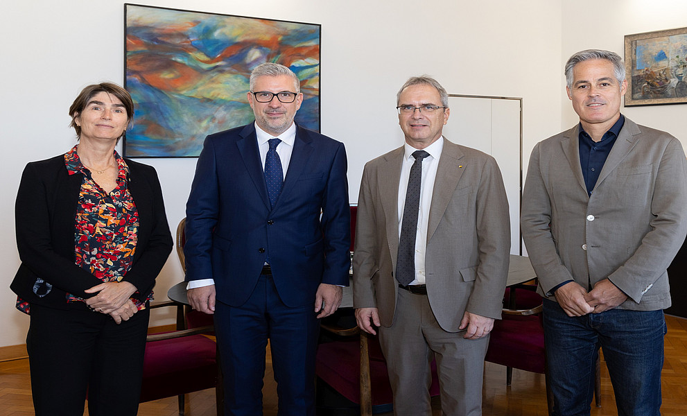 Vier Personen stehen nebeneinander in einem Raum mit Holzboden und weißen Wänden. Im Hintergrund sind zwei farbenfrohe Gemälde an der Wand zu sehen. Die Personen tragen formelle Kleidung, darunter Anzüge und Blazer. Hinter ihnen befinden sich rote Stühle und ein Tisch.