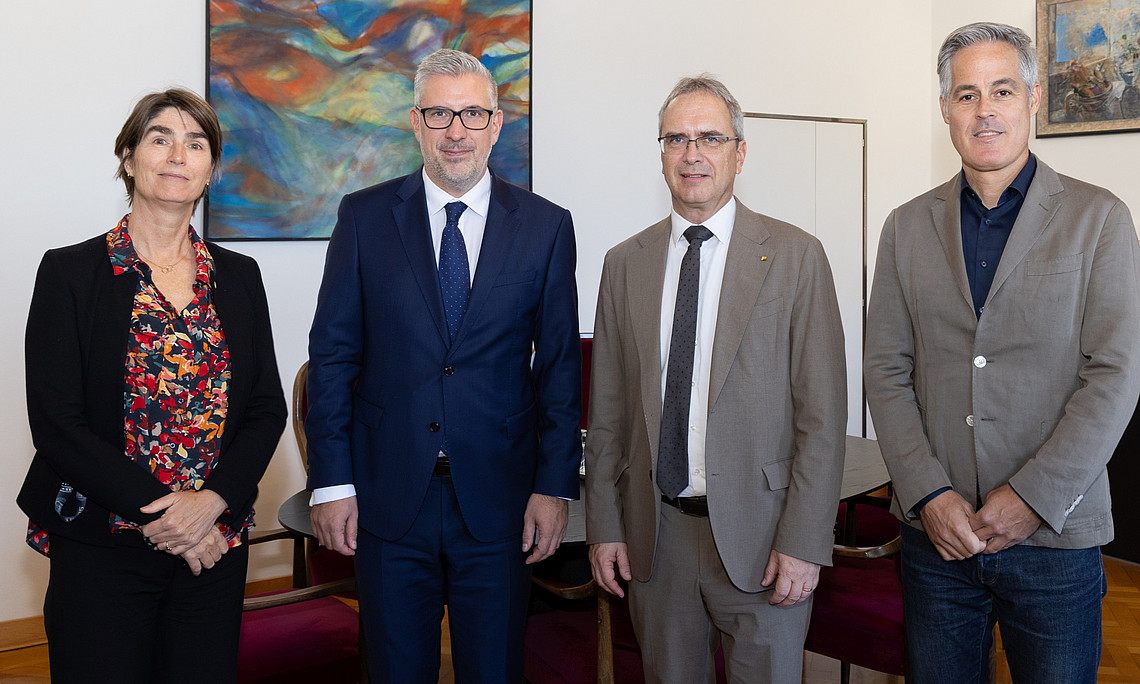 Vier Personen stehen nebeneinander in einem Raum mit Holzboden und weißen Wänden. Im Hintergrund sind zwei farbenfrohe Gemälde an der Wand zu sehen. Die Personen tragen formelle Kleidung, darunter Anzüge und Blazer. Hinter ihnen befinden sich rote Stühle und ein Tisch.