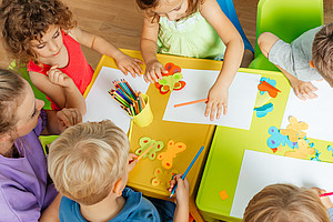 Lovely children glueing and cutting paper on colorful plastic tables. 