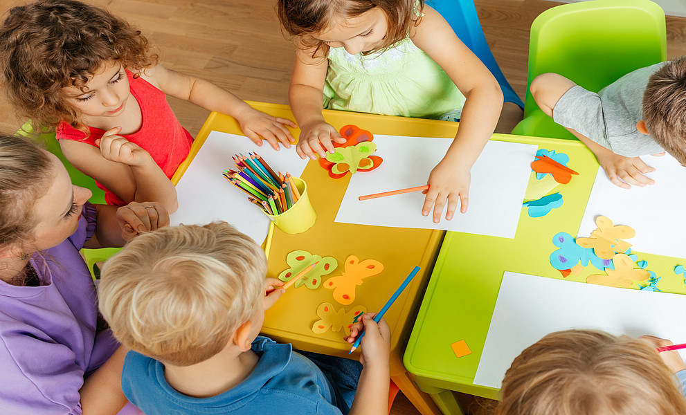 Niedliche Kinder kleben und schneiden Papier auf bunten Plastiktischen. 