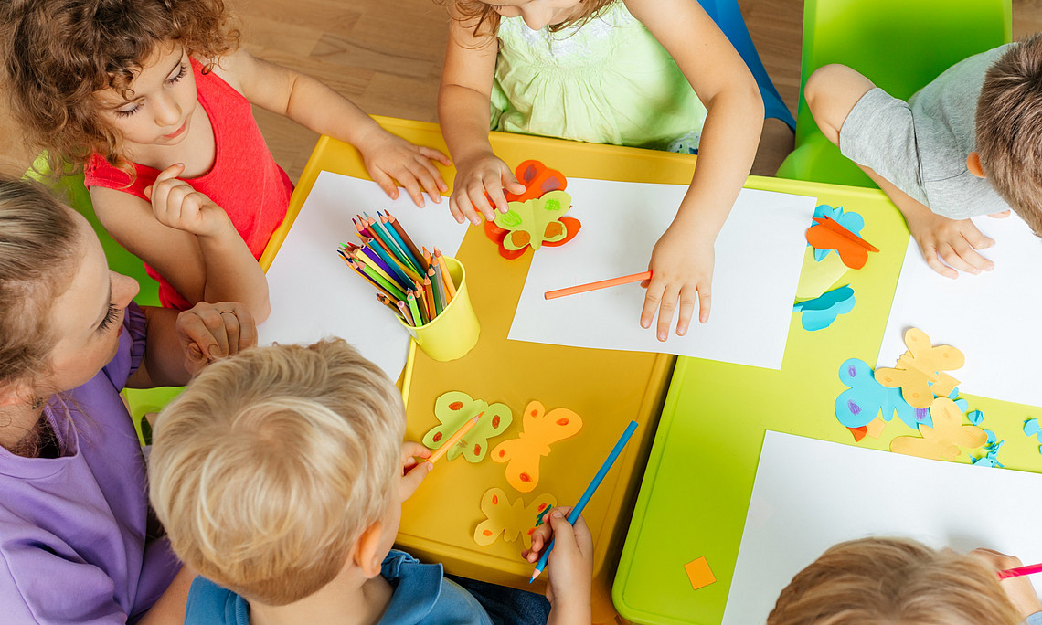 Lovely children glueing and cutting paper on colorful plastic tables. 