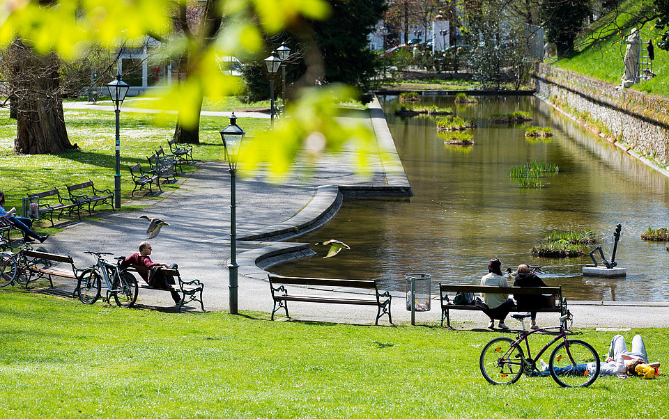 Ausschnitt des Stadtparks mit einem kleinen Gewässer, Grünflächen und vielen Bänken, worauf verteilt Menschen sitzen