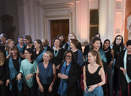 Ein Frauenchor, gekleidet in Schwarz und Blaugrün, singt in der Aula einer Universität mit Marmorstatuen im Hintergrund. ©Uni Graz/Schweiger