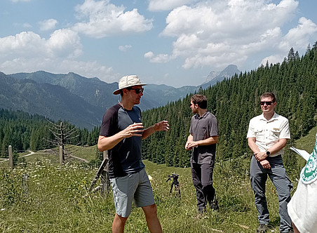 Ein Foto eines Teams in der Natur, auf einer Alm eines alpinen Berges mit grünen Wiesen und Fichten hinter ihnen. Die Gruppe steht um einen Mann, der graue Shorts, ein schwarzes T-Shirt und eine weiße Baseballmütze trägt, während er eine Sonnenbrille trägt und sich mit einer anderen Person unterhält, die ähnlich gekleidet ist, aber keine Brille trägt. 