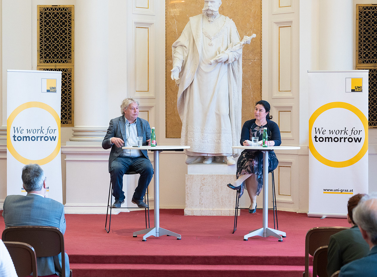 In einem prunkvollen Saal mit rotem Teppich sitzen zwei Personen auf hohen Hockern an Stehtischen auf einer kleinen Bühne. Hinter ihnen steht eine große weiße Statue in einer reich verzierten Nische. Links und rechts der Bühne stehen Roll‑Up‑Banner der **Uni Graz** mit dem gelben Kreis und dem Slogan **„We work for tomorrow“**. Im Vordergrund sitzen mehrere Zuhörer*innen auf Stühlen und verfolgen die Veranstaltung, die wie ein Vortrag oder eine Podiumsdiskussion wirkt. ©Uni Graz/Angele