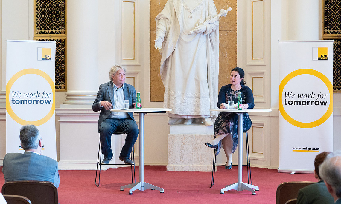 In a grand hall with a red carpet, two people are sitting on high stools at bar tables on a small stage. Behind them stands a large white statue in an ornately decorated niche. To the left and right of the stage are roll-up banners from **the University of Graz** featuring the yellow circle and the slogan **“We work for tomorrow”**. In the foreground, several members of the audience are seated on chairs, watching the event, which resembles a lecture or a panel discussion.