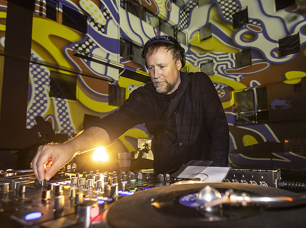 DJ Ocho ©Helmut Lunghammer A person behind a DJ desk playing music, a wall projection in the background ©Helmut Lunghammer