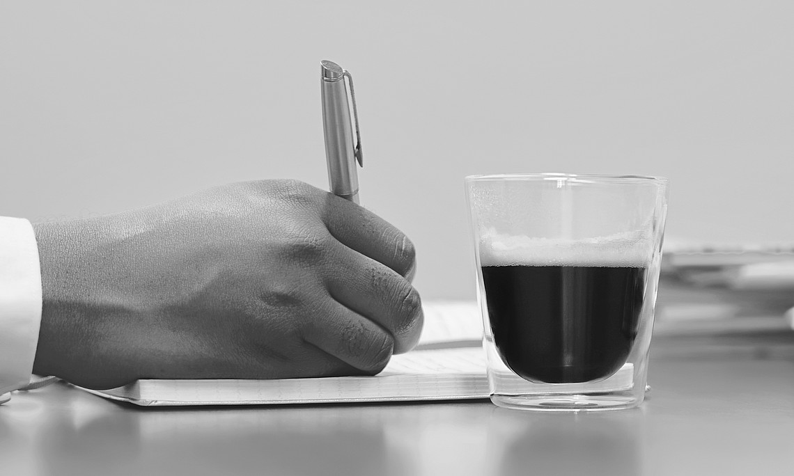 Eine Hand schreibt in einem Notizbuch, daneben steht eine Tasse.