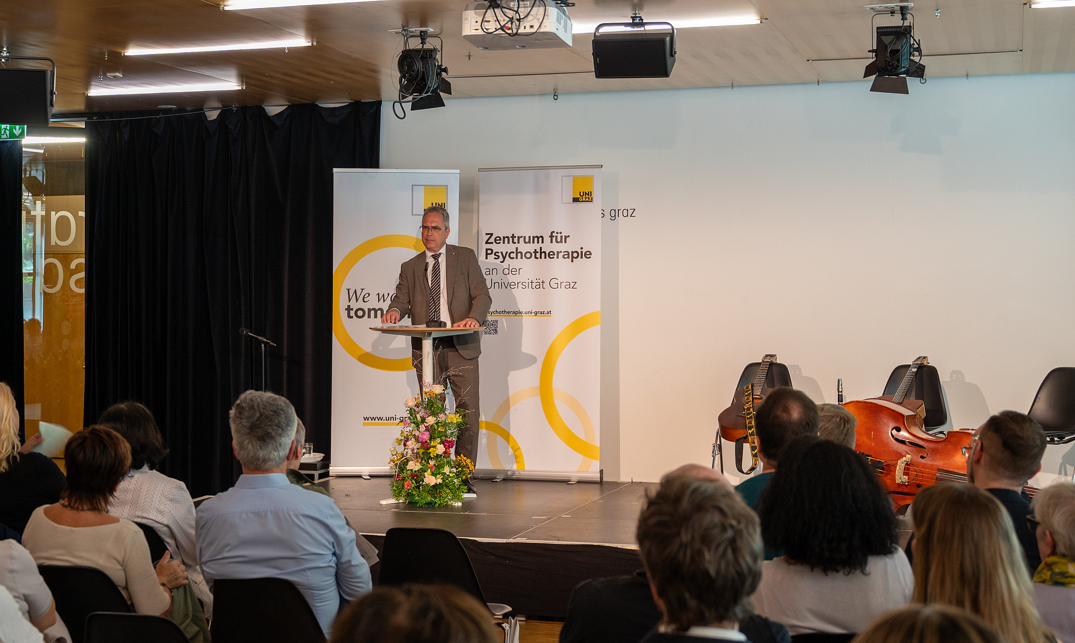 Mann mittleren Alters im Anzug spricht am Rednerpult vor Publikum, hinter ihm Banner „Zentrum für Psychotherapie“, rechts Musikinstrumente auf der Bühne. ©Uni Graz/Vilgut
