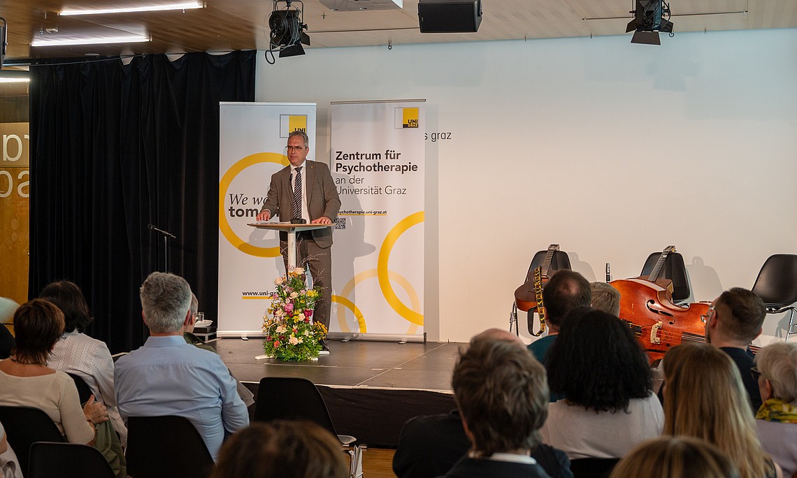 A middle-aged man in a suit is speaking at a lectern in front of an audience; behind him is a banner reading ‘Centre for Psychotherapy’; to the right, musical instruments are on the stage.
