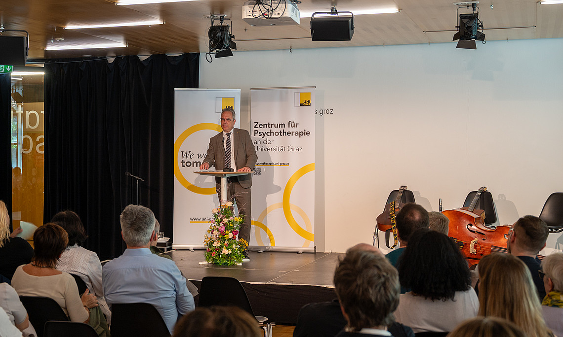 Mann mittleren Alters im Anzug spricht am Rednerpult vor Publikum, hinter ihm Banner „Zentrum für Psychotherapie“, rechts Musikinstrumente auf der Bühne.