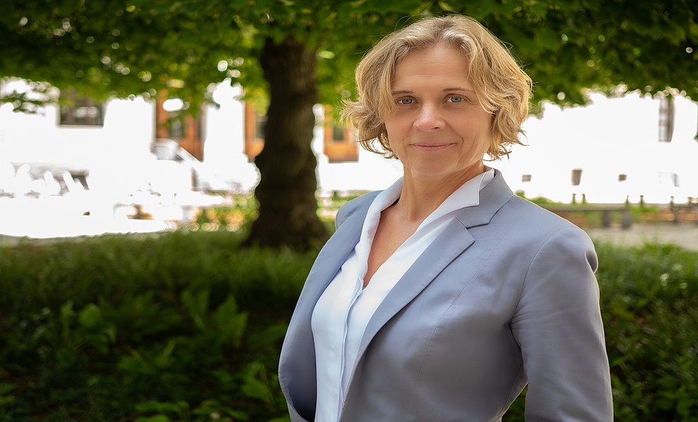 Eine Frau in einem grauen Blazer steht vor einem Baum mit grünem Laub