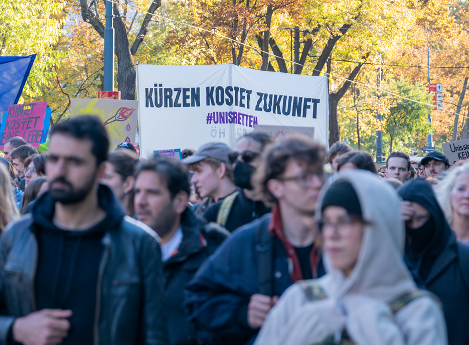 Menschenmenge bei einer Demonstration auf einer Straße; im Hintergrund ein Transparent mit der Aufschrift „Kürzen kostet Zukunft #UnisRetten“. 