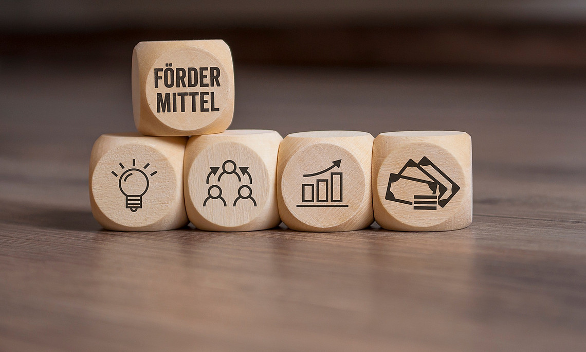 auf einem hölzernen Untergrund liegen fünf Würfel, auf deren Seiten zu sehen sind: eine Glühbirne, das Wort Fördermittel, drei Personen mit Bezugspfeilen, Geldscheine und größer werdende Balkendiagramm