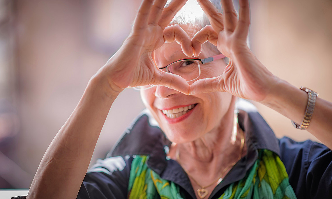 ältere Dame mit Brille macht mit ihren Fingern ein Herz und blickt durch