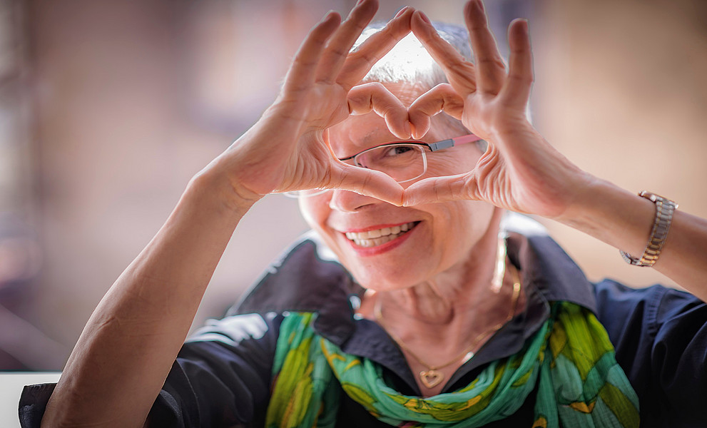 ältere Dame mit Brille macht mit ihren Fingern ein Herz und blickt durch