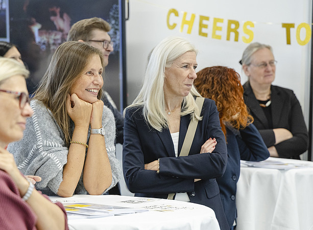  Gäste hören einer Präsentation zu; im Hintergrund steht der Schriftzug „CHEERS TO“. ©@foto-gasser.at