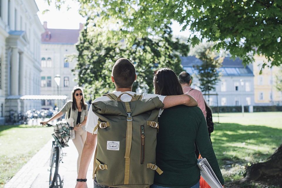 Zwei Studierende von hinten, die gemeinsam am Campus gehen, eine Person hat den Arm um die Freundin gelegt