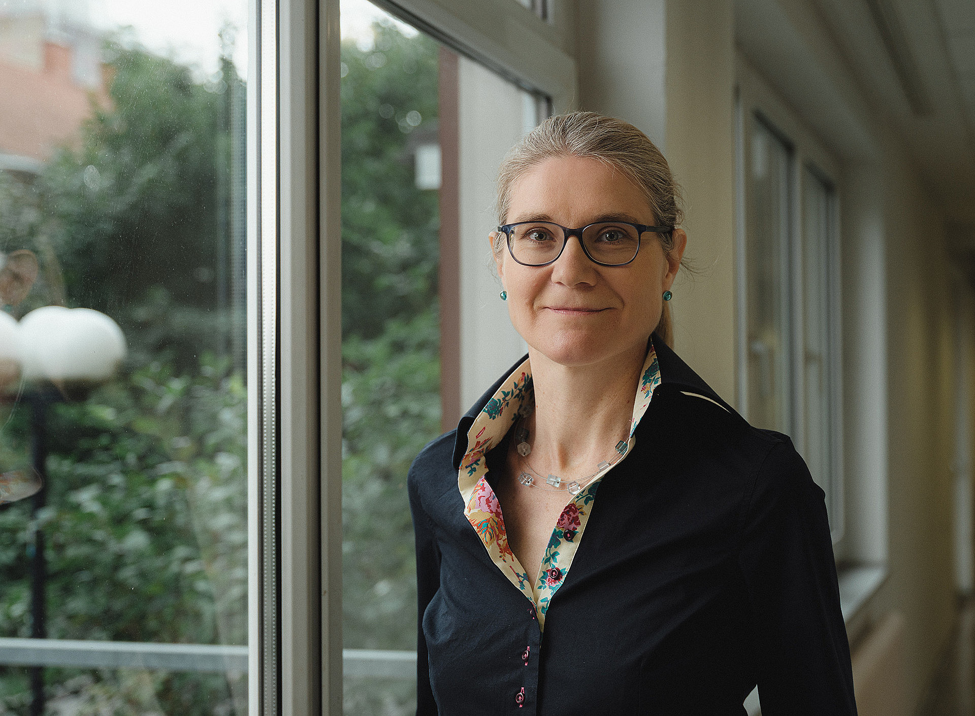 Porträt von Hermine Mitter, die in einem Gang vor einer Fensterfront steht. 