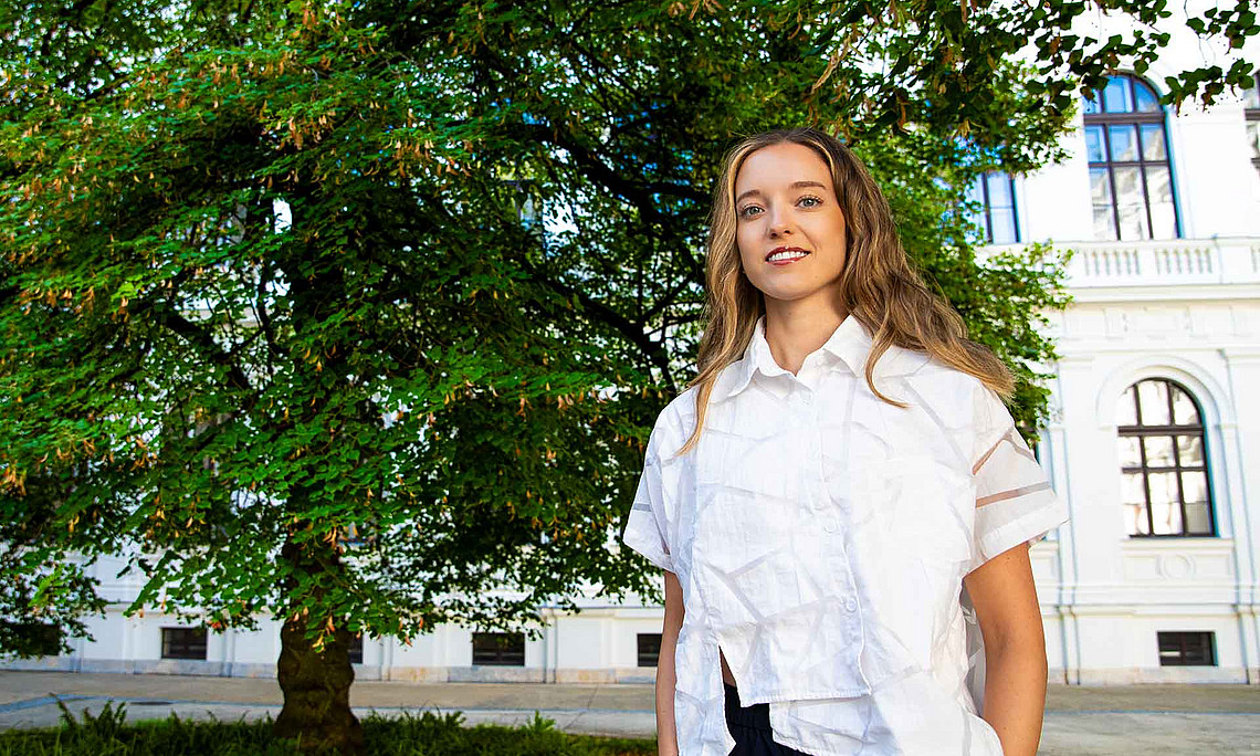 A young woman with long hair is standing in a white blouse and dark trousers in front of a large green tree and a historic building.