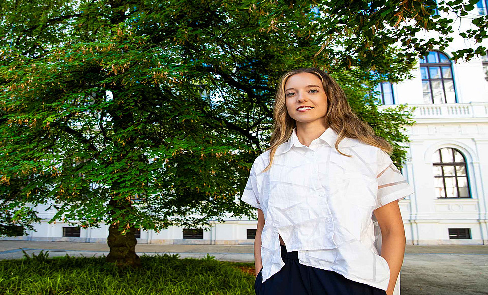 Junge Frau mit langen Haaren steht in weißer Bluse und dunkler Hose vor einem großen grünen Baum und einem historischen Gebäude.
