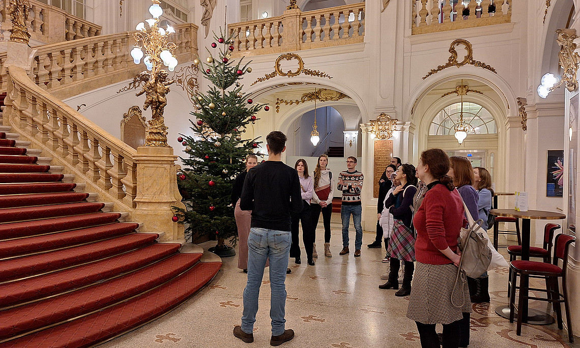 Eine Gruppe von Menschen steht im Foyer der Oper Graz, links ist eine rote Treppe mit prunkvollem Geländer.