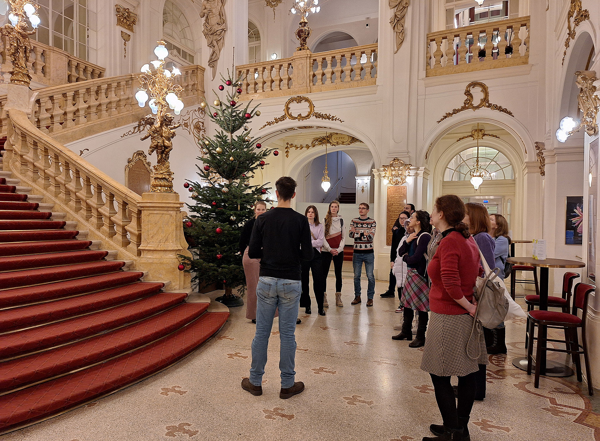 Eine Gruppe von Menschen steht im Foyer der Oper Graz, links ist eine rote Treppe mit prunkvollem Geländer. ©Uni Graz/Martina Mayer-Krauss