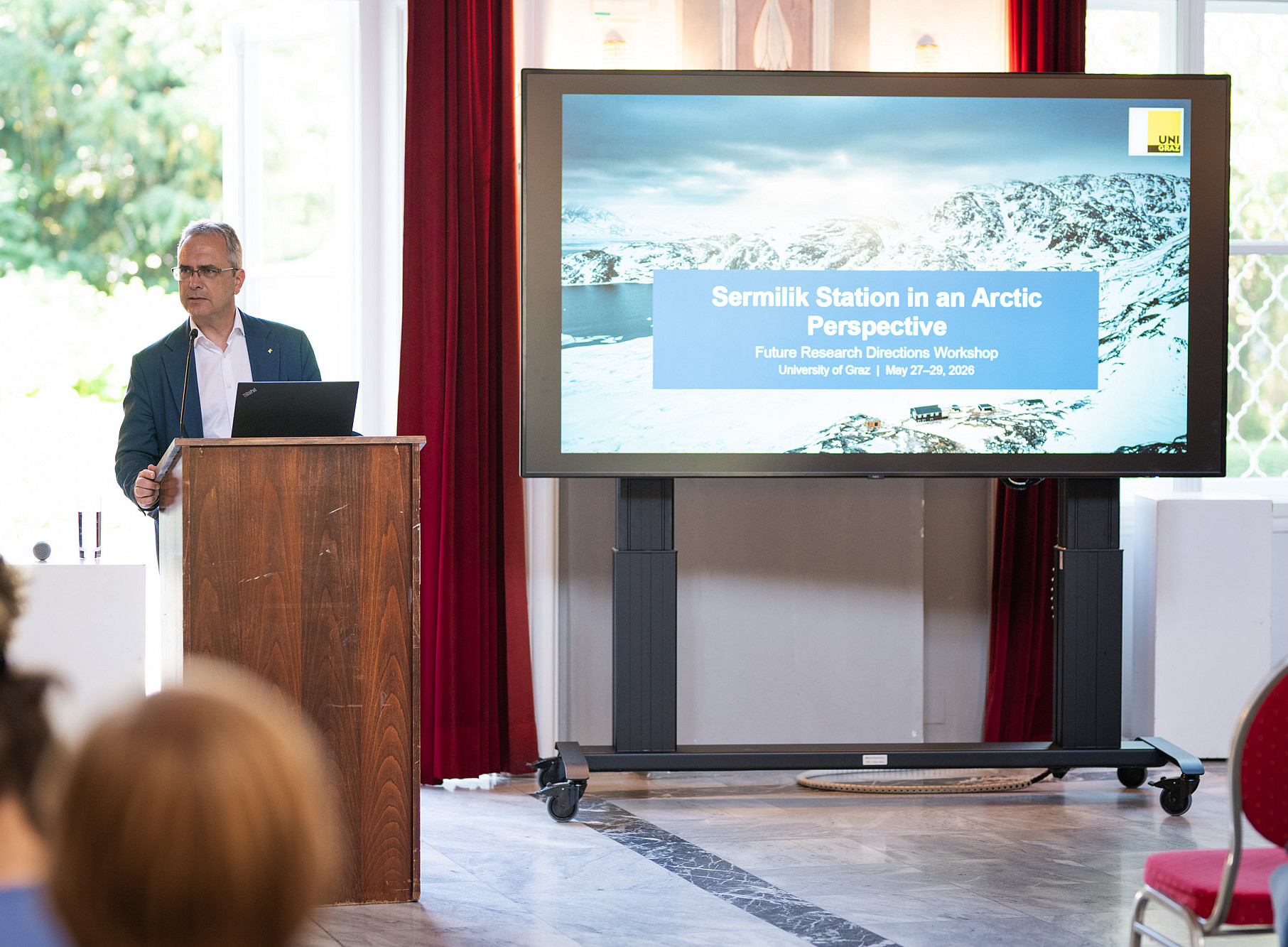 Eine Person im Anzug spricht zum Publikum; im Hintergrund ist ein großformatiges Bild von schneebedeckten Bergen und die Aufschrift „Sermilik Station in an Arctic Perspective“ zu sehen ©Uni Graz/Angele