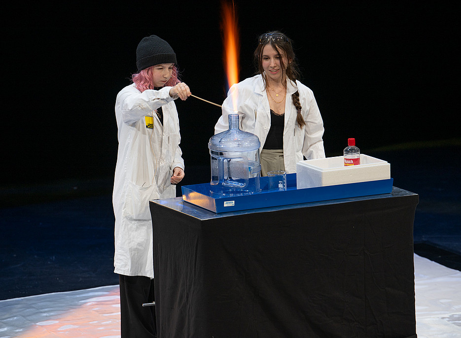 Zwei Personen in weißen Laborkitteln stehen hinter einem Tisch mit schwarzer Abdeckung, auf dem sich ein blauer Behälter mit einer großen transparenten Flasche, ein weißer Kasten und eine rote Flasche mit der Aufschrift „Wusch“ befinden; eine Person hält einen langen Stab, aus dem eine orangefarbene Flamme aufsteigt. 