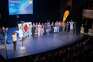 A group of people are standing next to each other on a stage, some are clapping, others are wearing red hats; on the left is a screen with the words “Chemical Life” and underneath it “The University of Graz's Chemical Christmas Show at the Schauspielhaus.” On the right is a yellow banner with the words “Studium an der Uni Graz”
