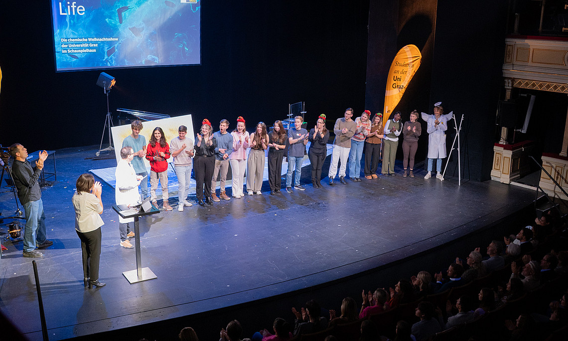 A group of people are standing next to each other on a stage, some are clapping, others are wearing red hats; on the left is a screen with the words “Chemical Life” and underneath it “The University of Graz's Chemical Christmas Show at the Schauspielhaus.” On the right is a yellow banner with the words “Studium an der Uni Graz”