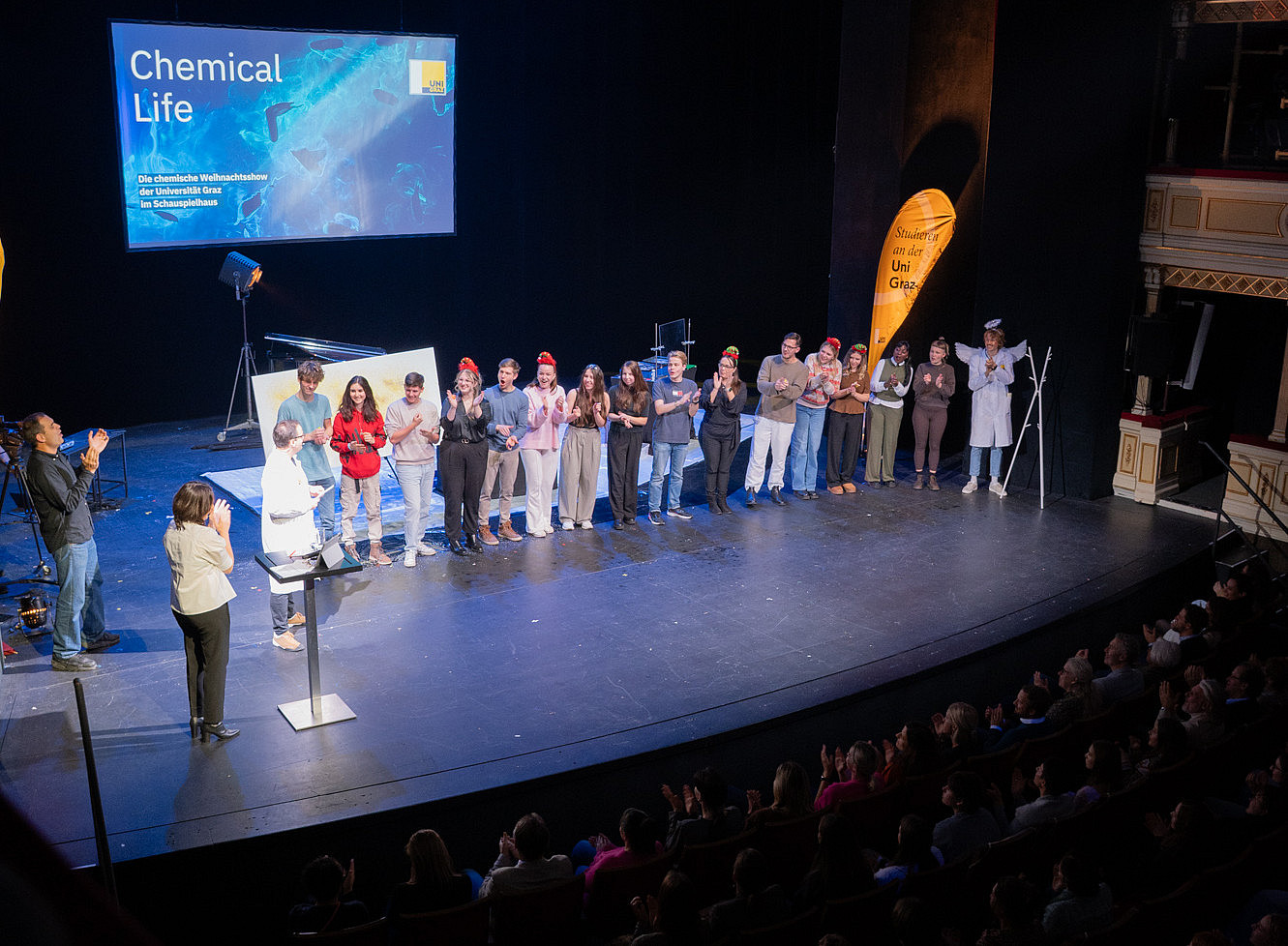 A group of people are standing next to each other on a stage, some are clapping, others are wearing red hats; on the left is a screen with the words “Chemical Life” and underneath it “The University of Graz's Chemical Christmas Show at the Schauspielhaus.” On the right is a yellow banner with the words “Studium an der Uni Graz” 