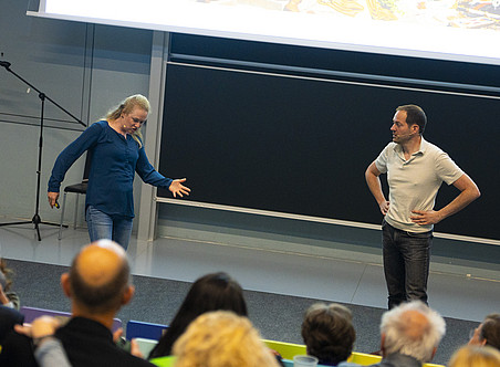 Ein Foto von zwei Personen vor einem Publikum, eine Frau und ein Mann, die stehen und lustige Dinge tun, während sie miteinander sprechen. Sie befinden sich in einem Hörsaal einer Universität mit einer Tafel im Hintergrund und sind beide leger gekleidet. ©Uni Graz/Nadja Fuchs